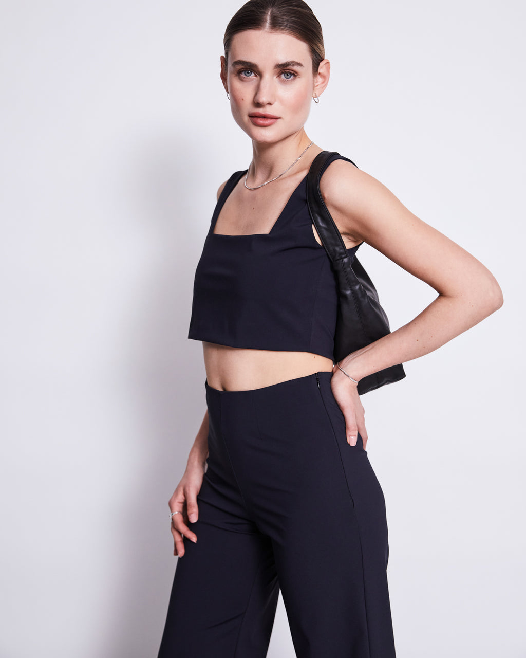 A woman with tied-back hair wears the jan-n-june CROP TOP CANCUN BLACK and matching high-waisted pants, holding a black shoulder bag, standing against a plain white background.