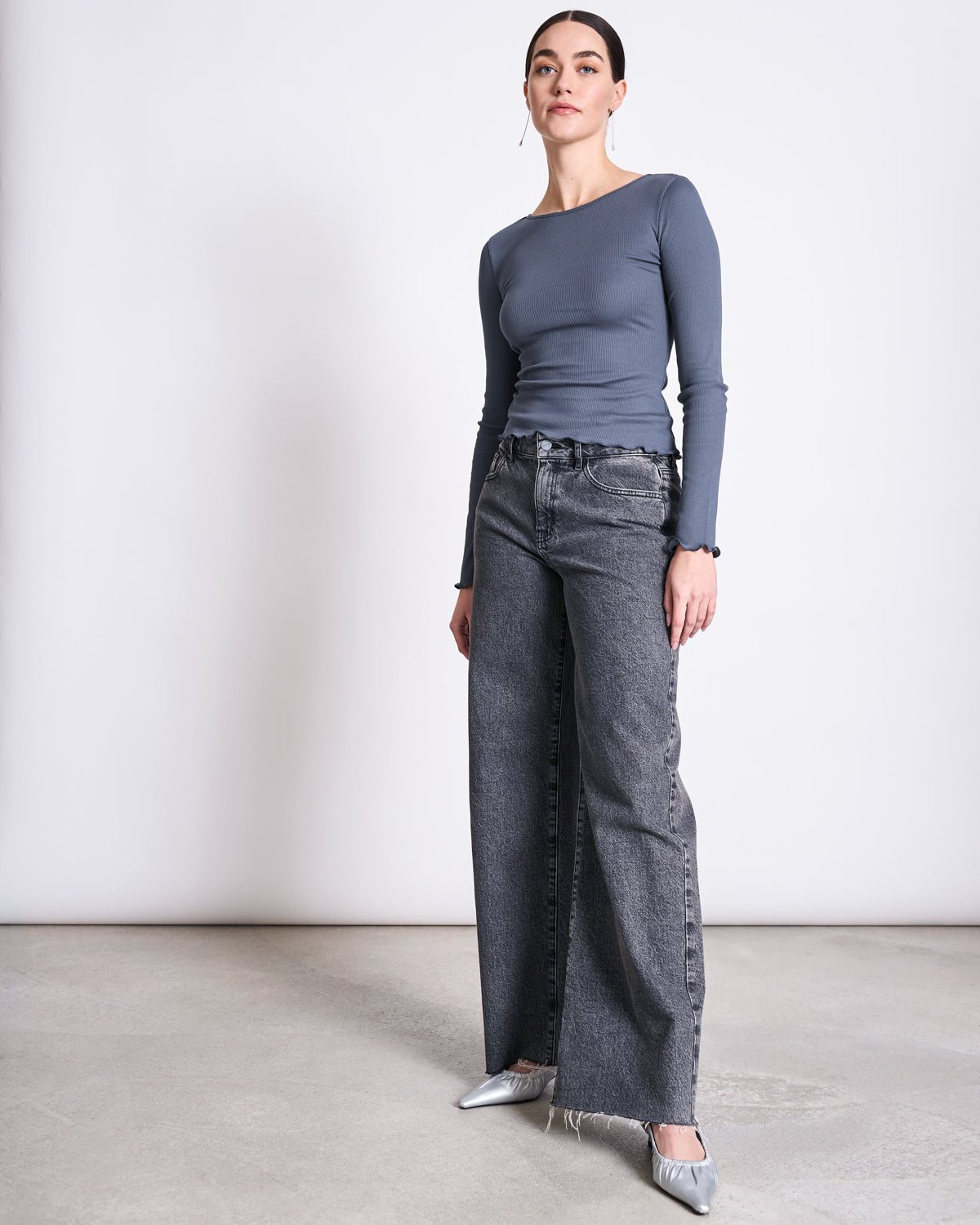 A woman with her hair tied back and a neutral expression stands against a plain background, wearing the jan-n-june MINIRIB LONGSLEEVE AVERSA TURBULENCE GOTS blue crop top, high-waisted wide-leg gray jeans, and silver pointed-toe shoes.