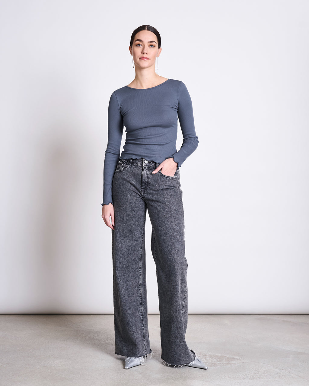 A woman stands against a plain background wearing the jan-n-june MINIRIB LONGSLEEVE AVERSA TURBULENCE GOTS top, paired with high-waisted wide-leg gray jeans and pointed-toe shoes. Her hair is pulled back, and one hand is in her pocket.