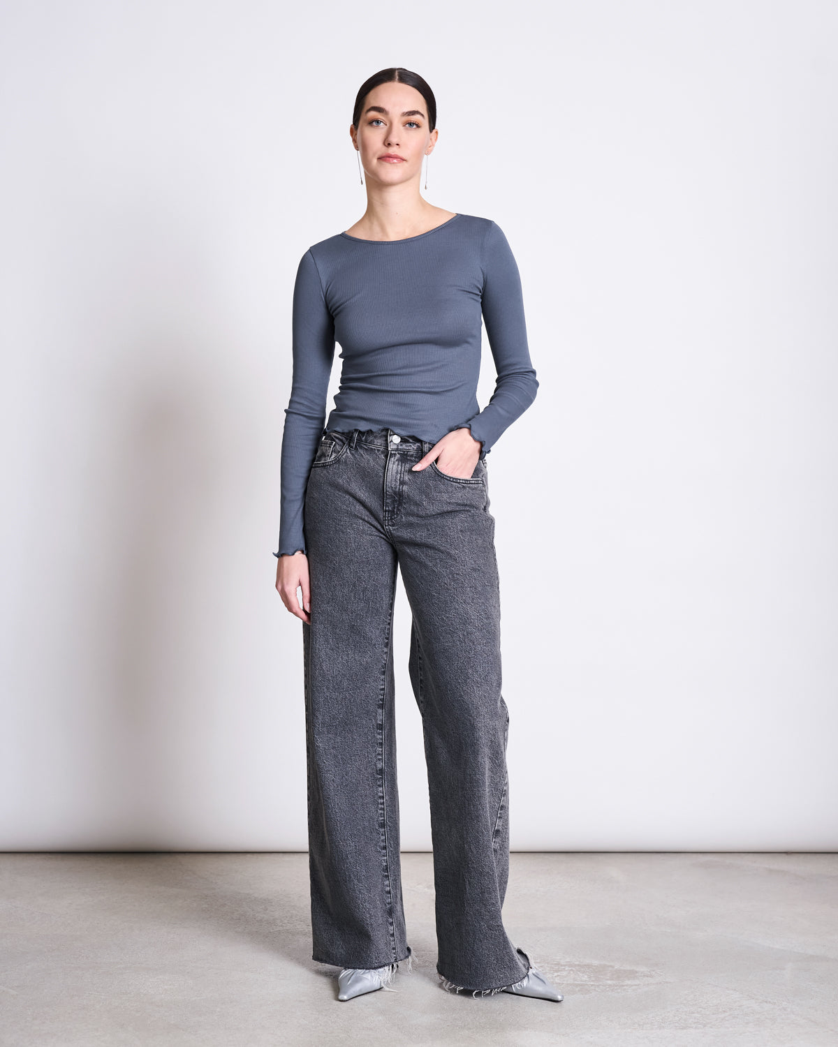 A woman stands against a plain background wearing the jan-n-june MINIRIB LONGSLEEVE AVERSA TURBULENCE GOTS top, paired with high-waisted wide-leg gray jeans and pointed-toe shoes. Her hair is pulled back, and one hand is in her pocket.