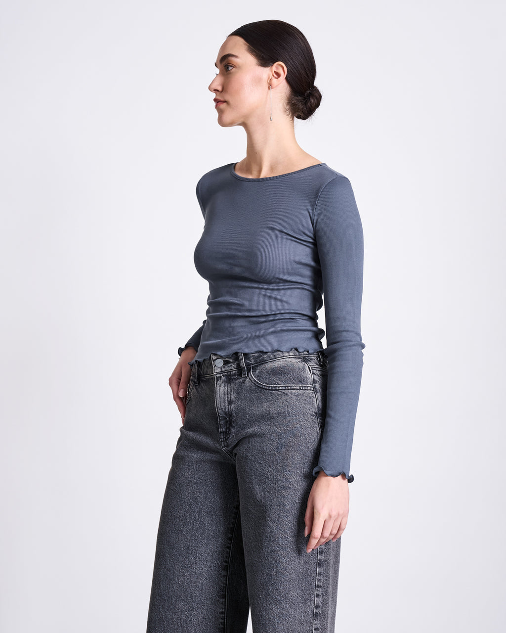 A woman with dark hair in a low bun wears the jan-n-june MINIRIB LONGSLEEVE AVERSA TURBULENCE GOTS and high-waisted, loose gray jeans. She stands sideways with one hand in her pocket, looking ahead against a plain background.