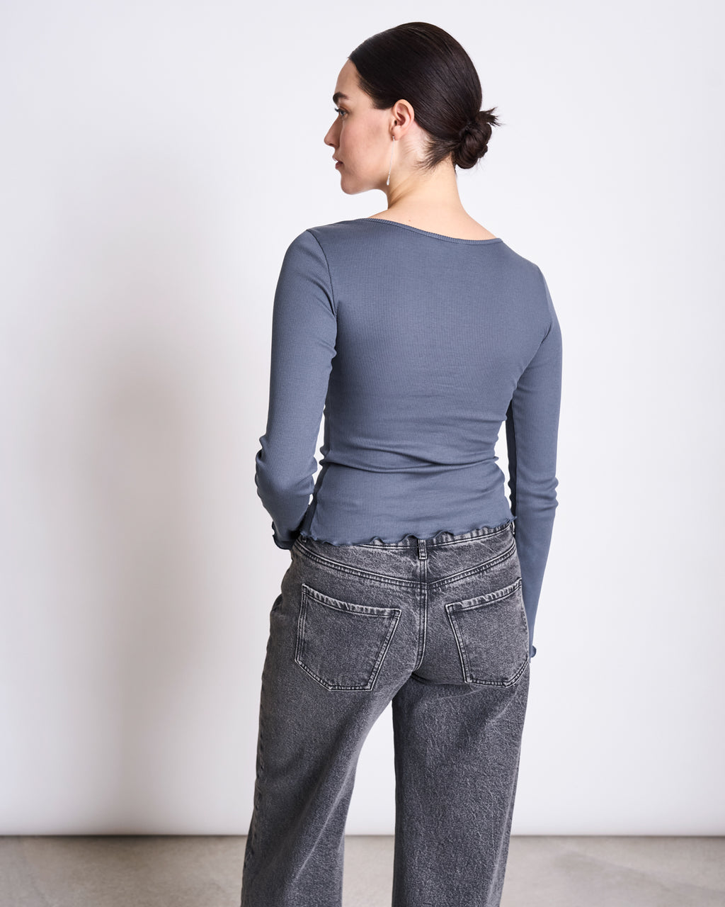 A person with dark hair in a low bun faces away from the camera, wearing jan-n-junes MINIRIB LONGSLEEVE AVERSA TURBULENCE GOTS, a slim-fit gray organic cotton rib top, paired with high-waisted faded black jeans against a plain light background.