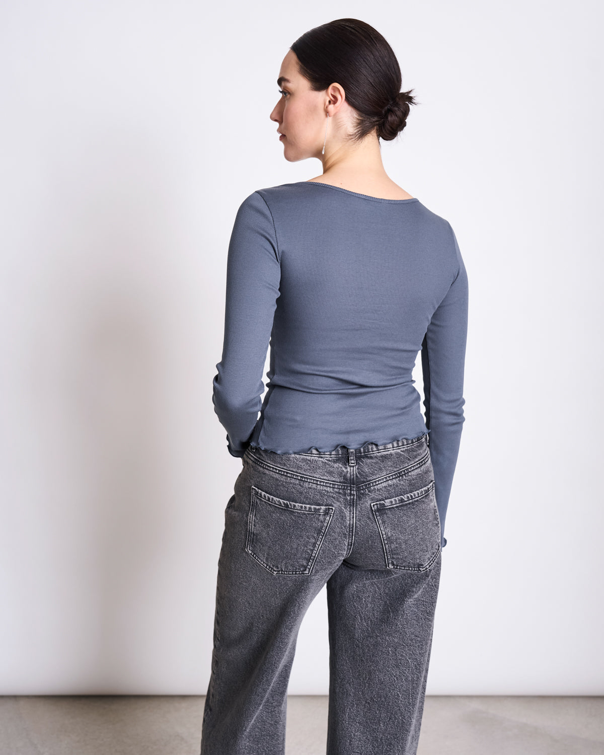 A person with dark hair in a low bun faces away from the camera, wearing jan-n-junes MINIRIB LONGSLEEVE AVERSA TURBULENCE GOTS, a slim-fit gray organic cotton rib top, paired with high-waisted faded black jeans against a plain light background.