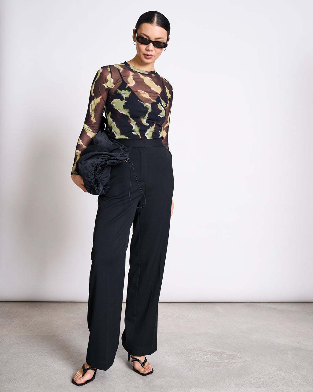 A woman stands against a plain background wearing jan-n-june’s MESH LONGSLEEVE DORA MOSSFIRE top, black sunglasses, high-waisted black pants, black heeled sandals, and holding a black ruched bag.