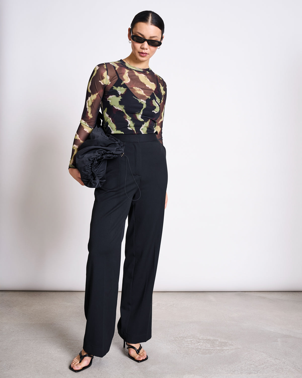 A woman stands against a plain background wearing jan-n-june’s MESH LONGSLEEVE DORA MOSSFIRE top, black sunglasses, high-waisted black pants, black heeled sandals, and holding a black ruched bag.