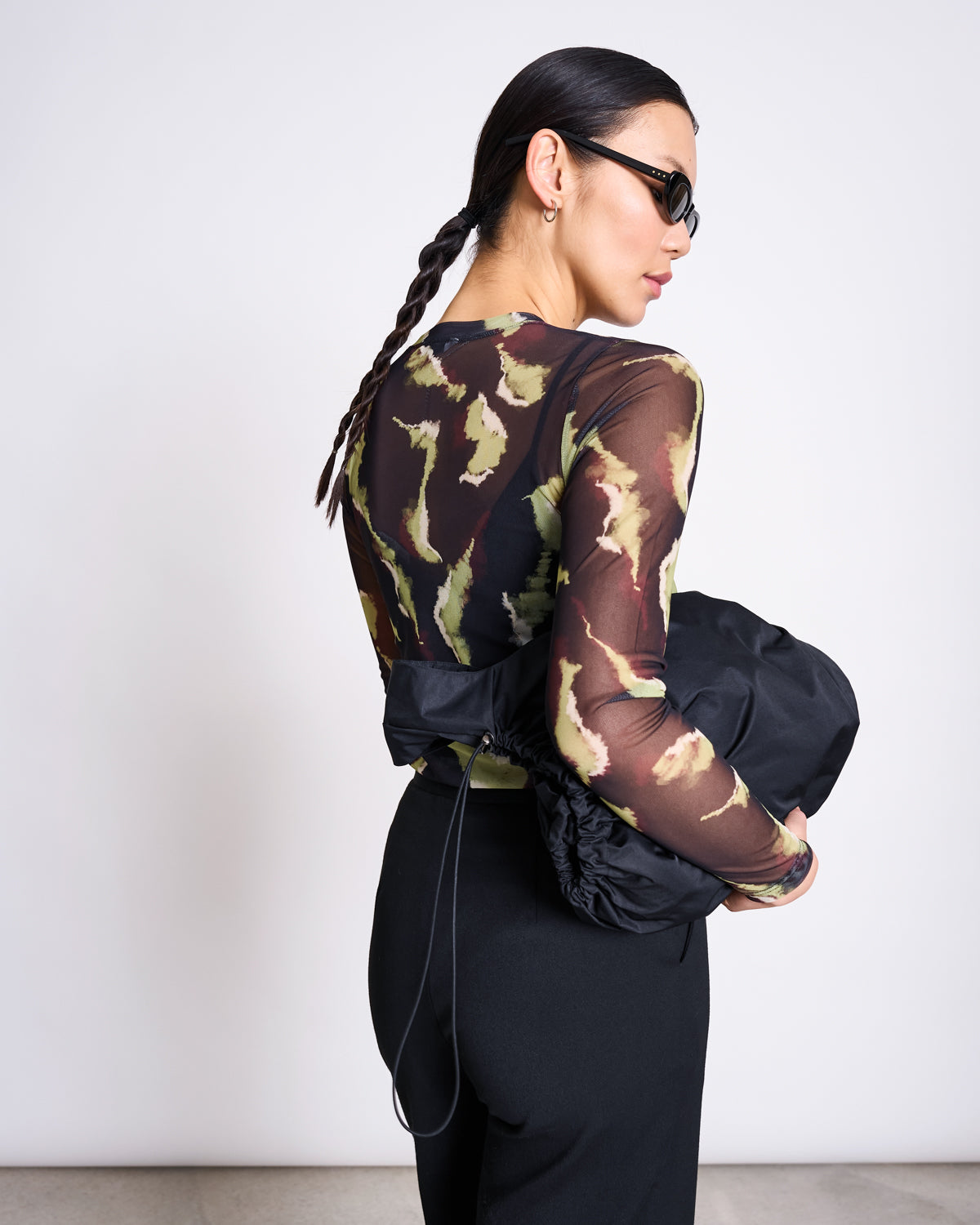 A woman in sunglasses and a braided ponytail faces away, modeling the jan-n-june MESH LONGSLEEVE DORA MOSSFIRE top. She holds a black bag at her side, standing before a plain white wall.