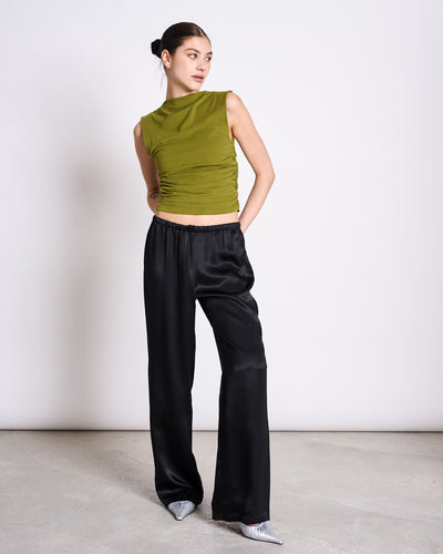 A woman stands against a plain background wearing the jan-n-june RUFFLE TOP RAFO WOODY OLIVE GOTS, paired with loose black satin pants and silver pointed-toe shoes. Her hair is pulled back, and one hand is in her pocket.