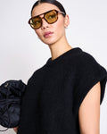 A woman with slicked-back hair wears large yellow-tinted sunglasses and the jan-n-june WOOLY SLIPOVER THANDA BLACK sweater, holding a black ruched bag against a plain light background.
