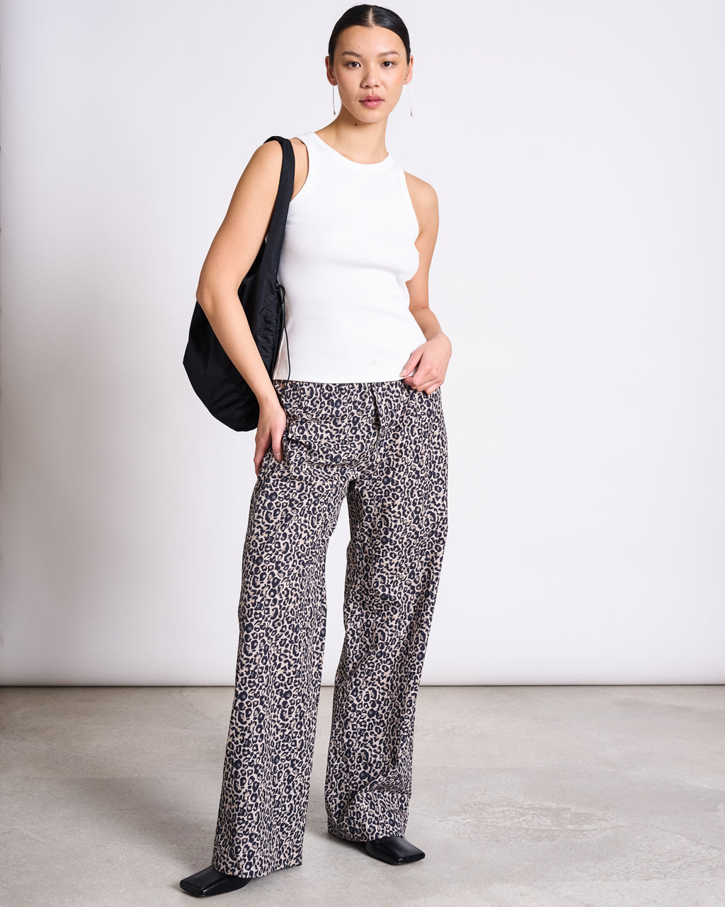 A woman with her hair tied back stands against a plain background, wearing jan-n-junes COTTON PANTS SOBY LEO PRINT, a white sleeveless top, black shoes, and a black shoulder bag. She has a neutral expression.