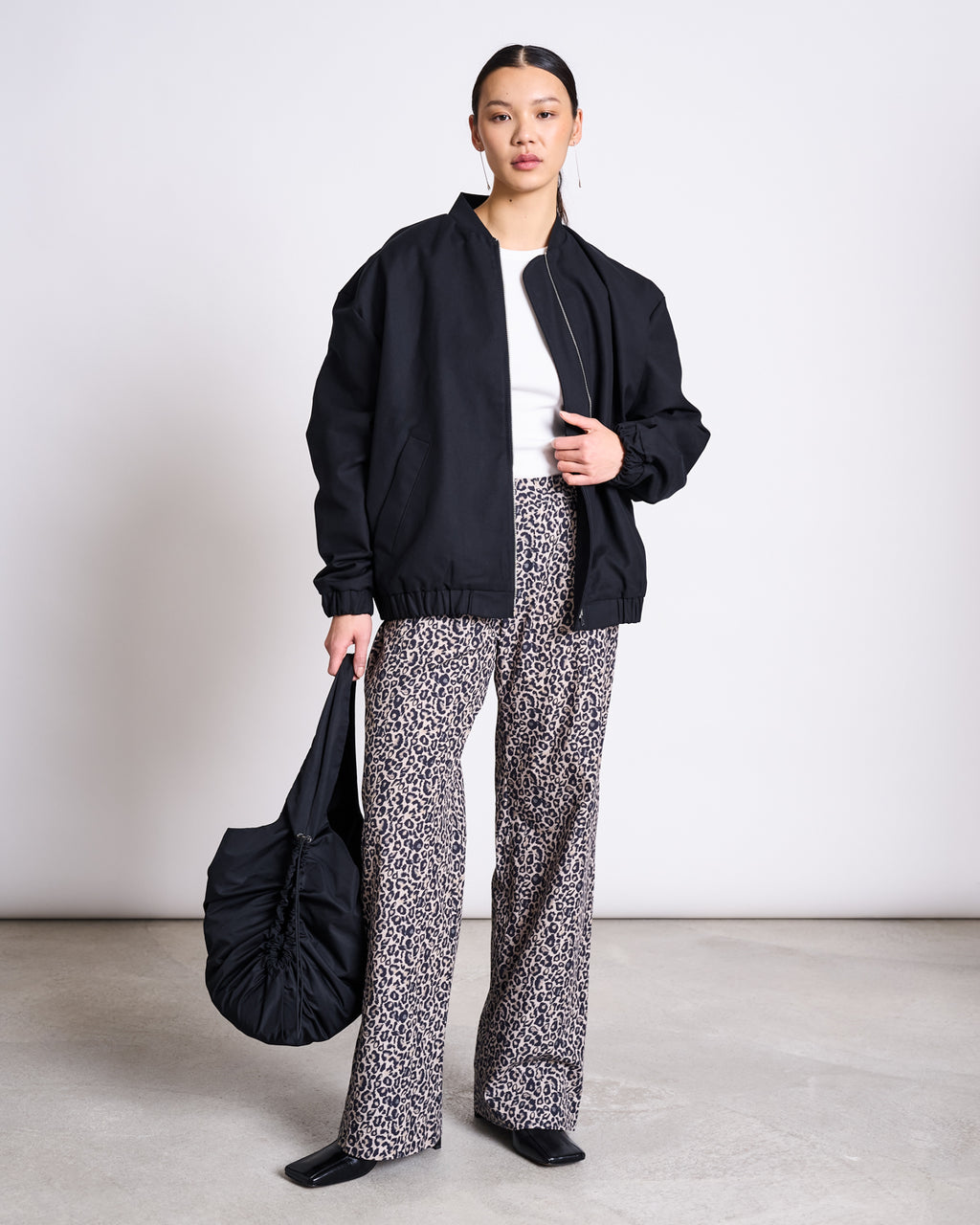 A woman with her hair tied back poses indoors against a light backdrop, wearing jan-n-junes COTTON PANTS SOBY LEO PRINT, a black bomber jacket over a white top, black square-toe shoes, and holding a black ruched handbag.