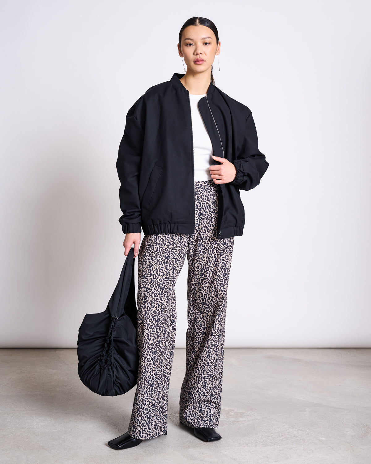 A woman with her hair tied back poses indoors against a light backdrop, wearing jan-n-junes COTTON PANTS SOBY LEO PRINT, a black bomber jacket over a white top, black square-toe shoes, and holding a black ruched handbag.