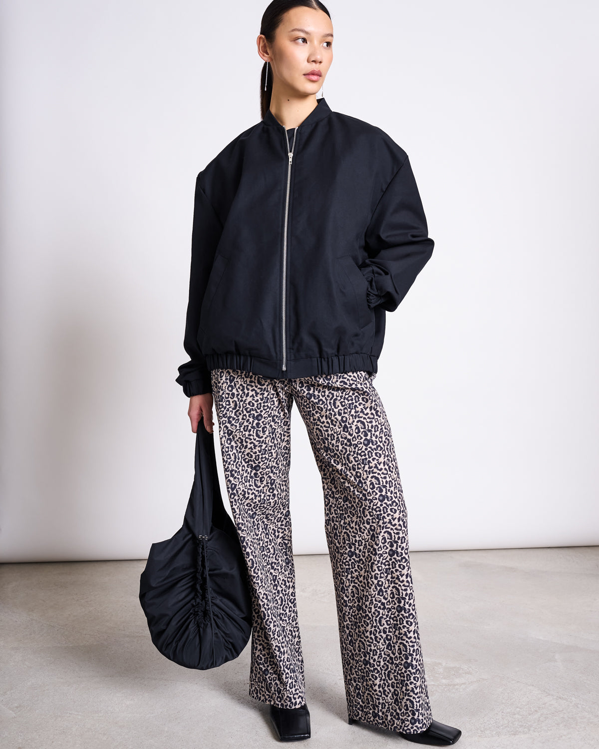 A woman stands against a plain background wearing wide-leg COTTON PANTS SOBY LEO PRINT by jan-n-june, a black oversized zip-up jacket, black ankle boots, and holding a black ruched bag.