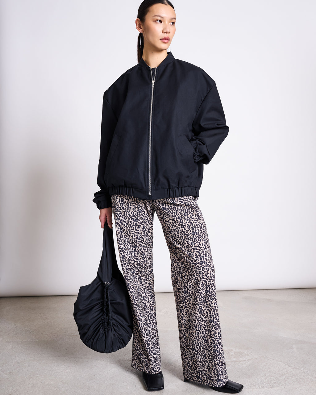 A woman stands against a plain background wearing the jan-n-june OVERSIZED BLOUSON TANARO BLACK, leopard print wide-leg pants, and black shoes. She holds a black ruched bag, looking to the side with her hair pulled back.