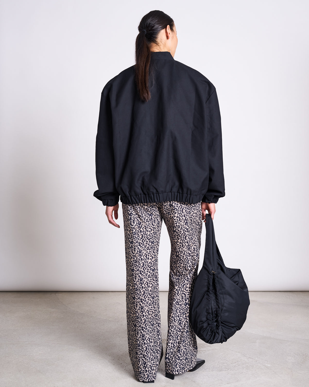A person stands facing away, wearing jan-n-junes COTTON PANTS SOBY LEO PRINT with an oversized black jacket and black shoes, holding a large black bag in their right hand against a light, plain background.