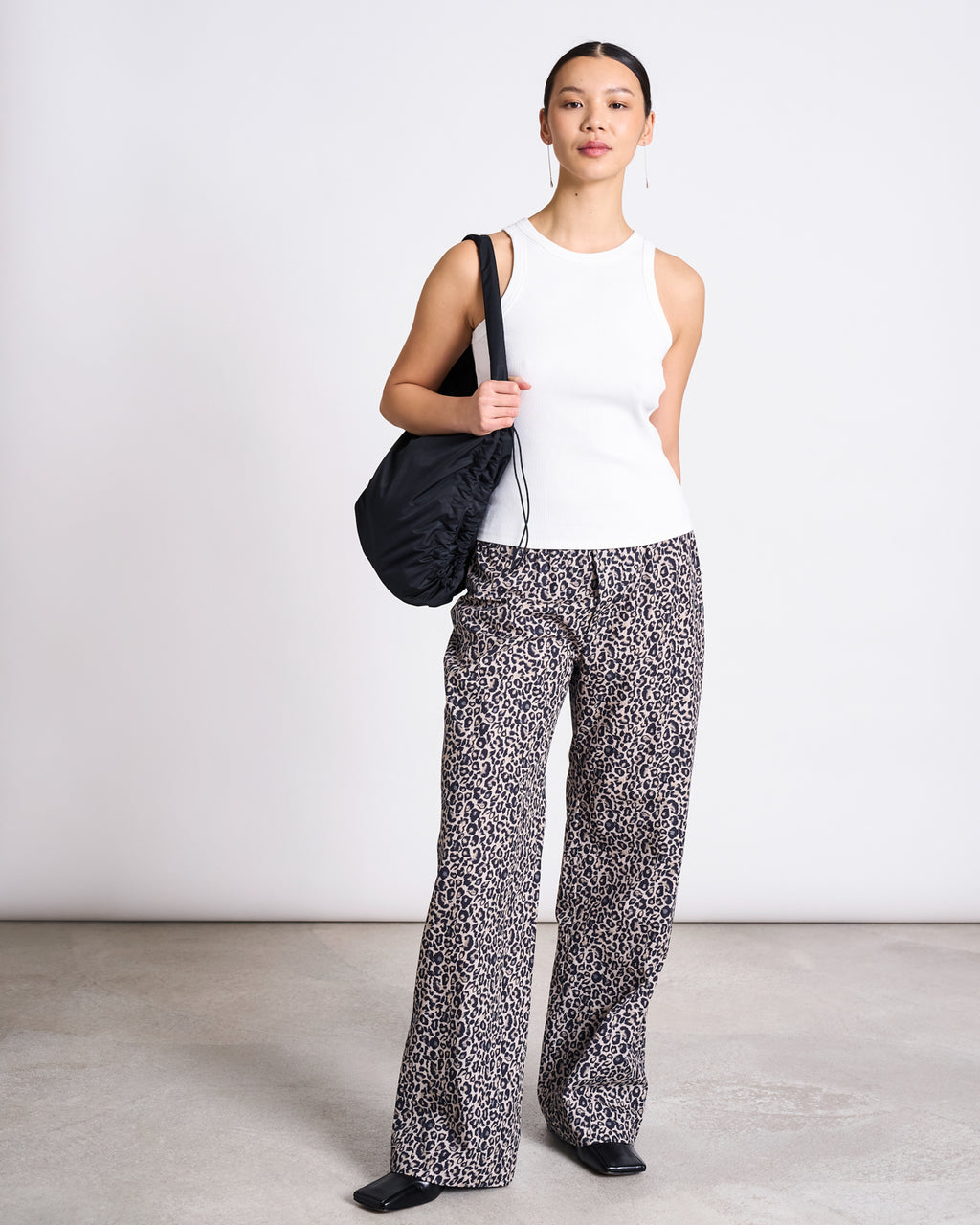 A woman stands indoors against a plain wall, wearing jan-n-junes COTTON PANTS SOBY LEO PRINT with a white sleeveless top, black shoes, and a black shoulder bag. She looks forward with a neutral expression.