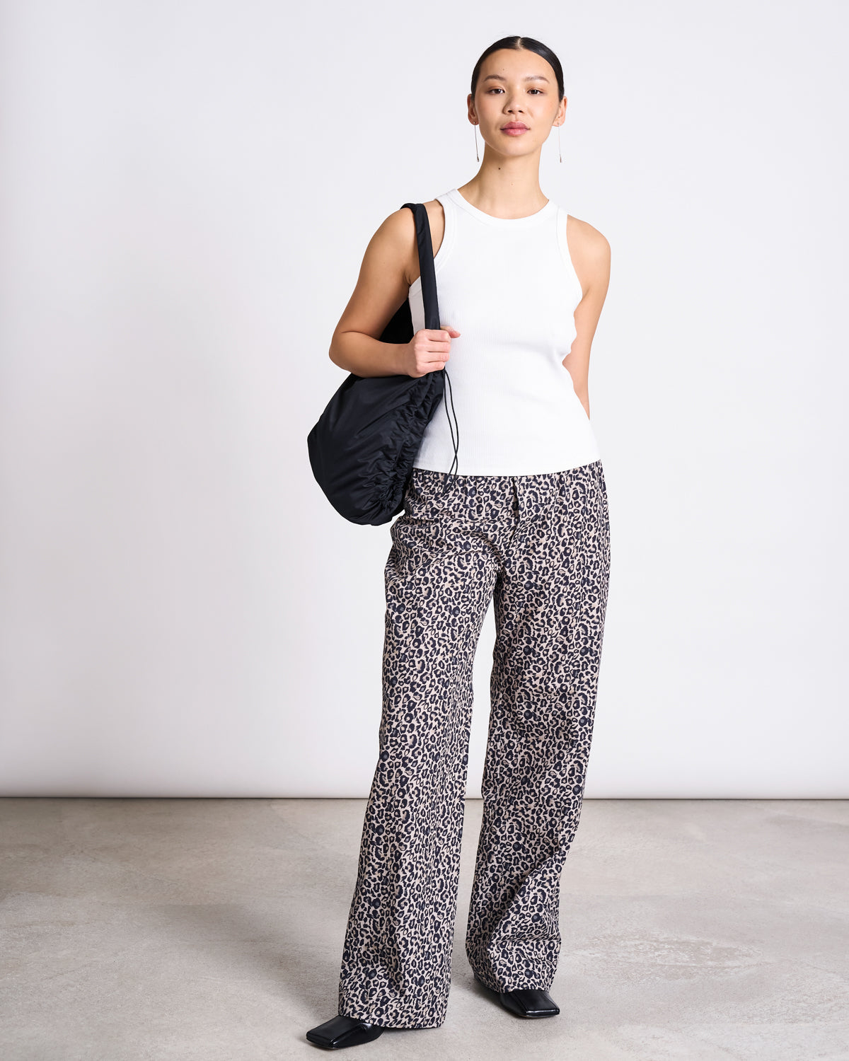 A woman stands indoors against a plain wall, wearing jan-n-junes COTTON PANTS SOBY LEO PRINT with a white sleeveless top, black shoes, and a black shoulder bag. She looks forward with a neutral expression.