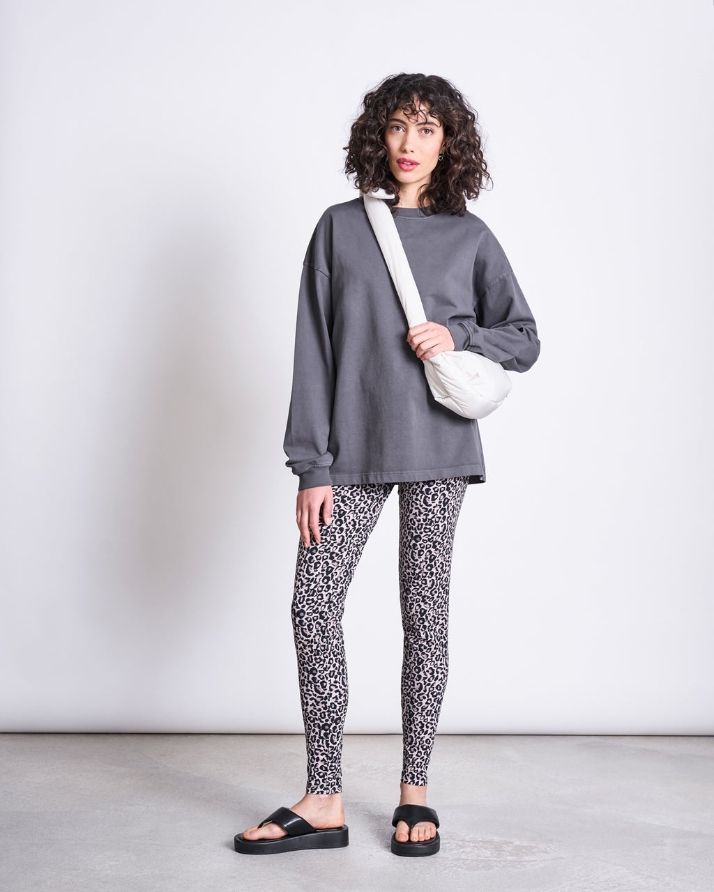 A woman with curly hair wears jan-n-june LEGGINGS LEO GOTS, a loose dark gray sweatshirt, black slide sandals, and a white crossbody bag, standing against a plain light background.