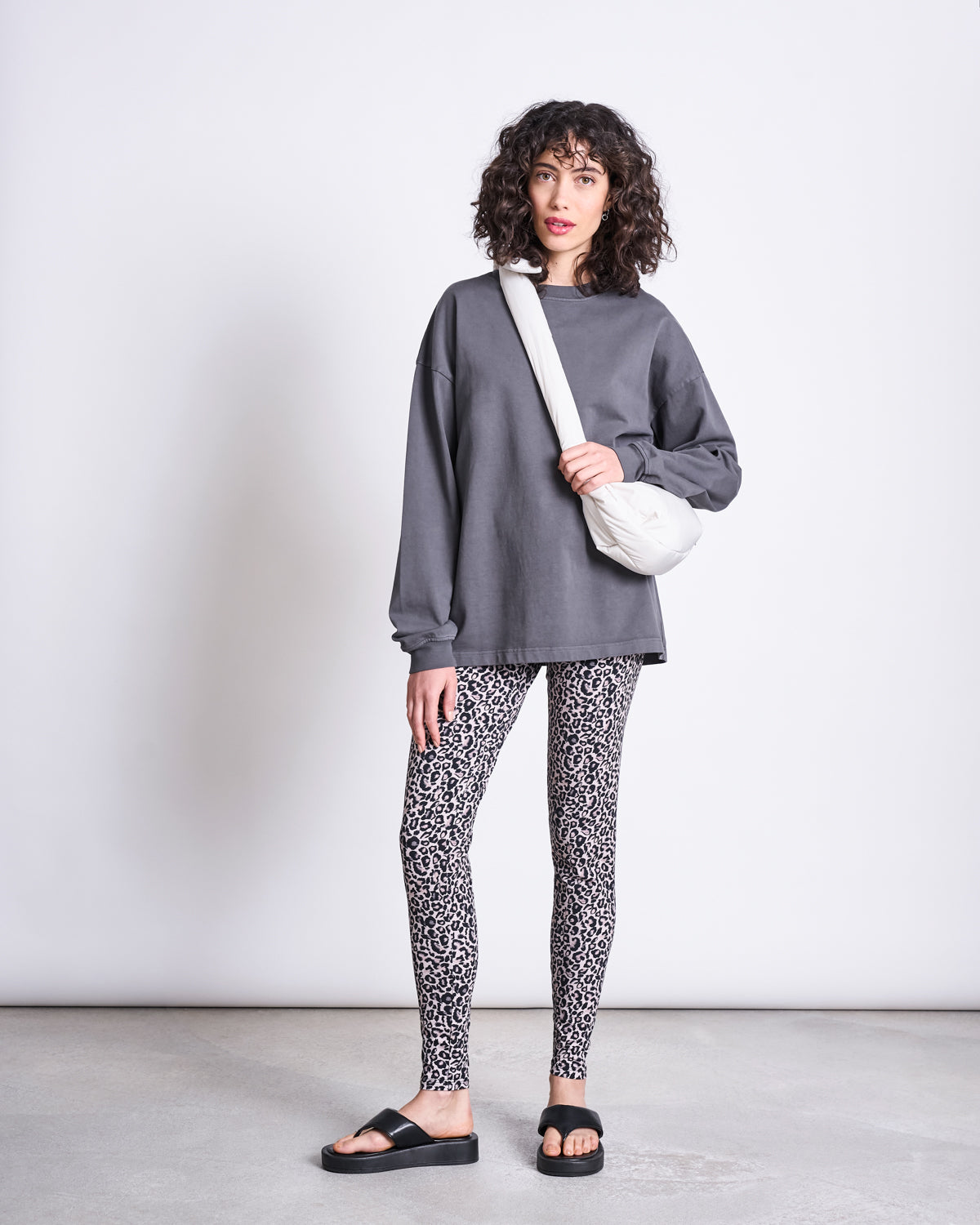 A woman with curly hair wears jan-n-june LEGGINGS LEO GOTS, a loose dark gray sweatshirt, black slide sandals, and a white crossbody bag, standing against a plain light background.