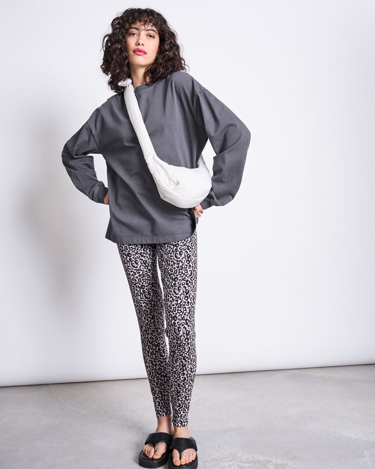 A woman poses in a minimal studio wearing a loose gray top, leopard print leggings, black slide sandals, and the jan-n-june BAG BIWA PUFFY MICRO ICE—an ultralightweight recycled polyester bag. She has curly hair and one hand on her hip.