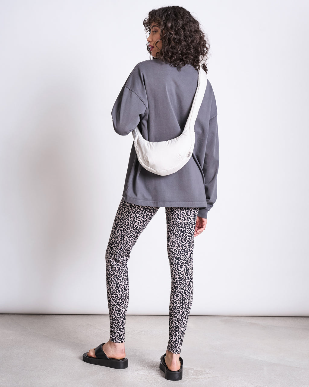 A woman with curly hair stands facing away, wearing jan-n-june’s LEGGINGS LEO GOTS, a loose gray long-sleeve shirt, black sandals, and a white crossbody bag, against a plain white wall and concrete floor.