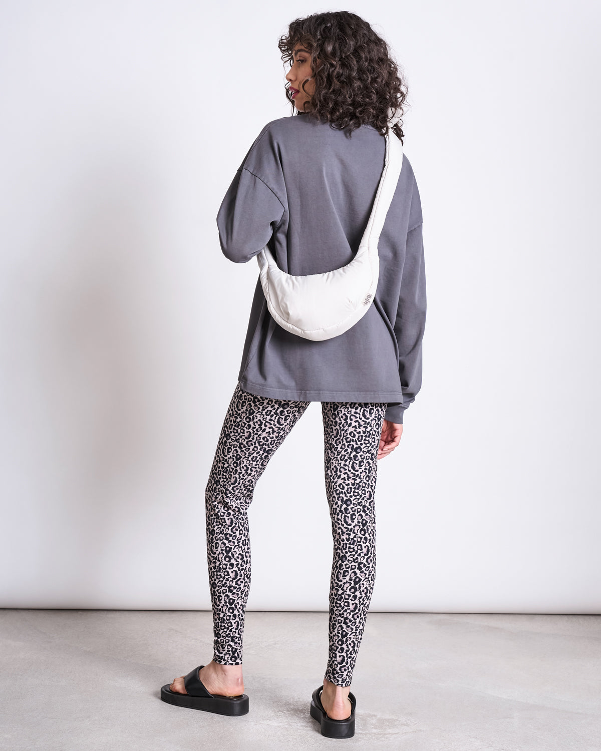 A woman with curly hair stands facing away, wearing jan-n-june’s LEGGINGS LEO GOTS, a loose gray long-sleeve shirt, black sandals, and a white crossbody bag, against a plain white wall and concrete floor.