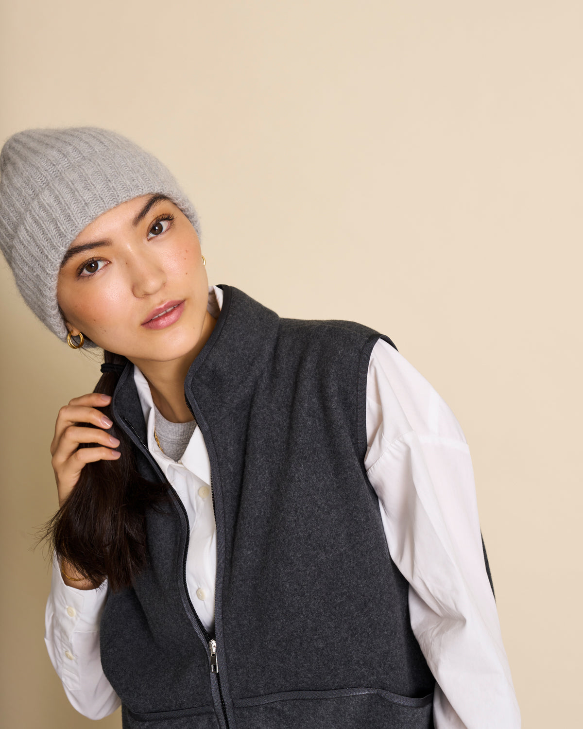 A woman wears the jan-n-june WOOLY KNIT BEANIE BJON LIGHT GREY, a white button-up shirt, and a dark gray zip-up vest, looking at the camera against a beige background while holding her hair.