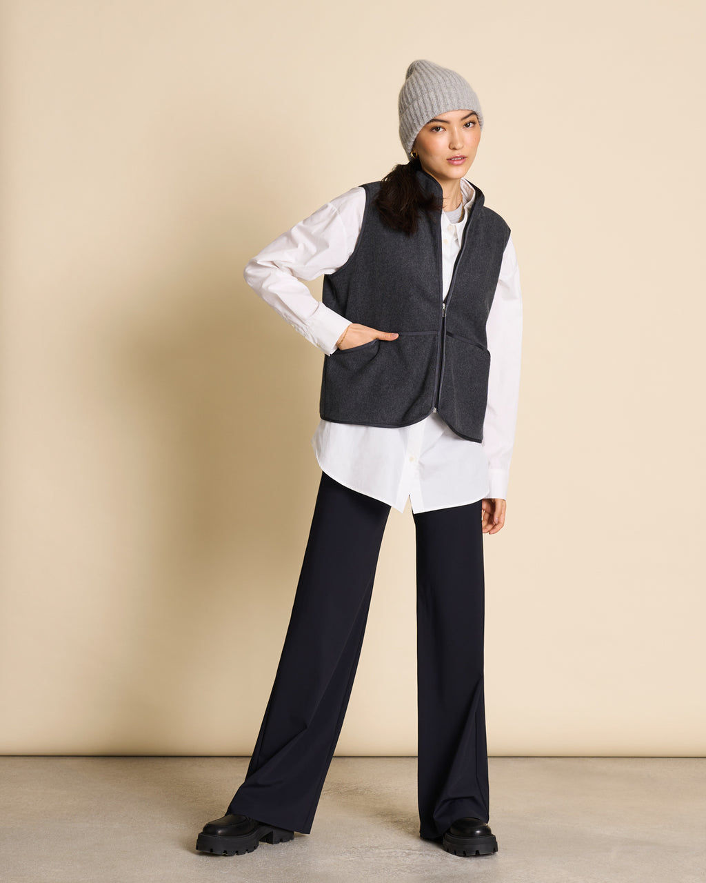 A woman wearing the jan-n-june WOOLY KNIT BEANIE BJON LIGHT GREY, a dark gray vest, white shirt, and black wide-leg pants stands against a beige background, looking at the camera with one hand on her hip.