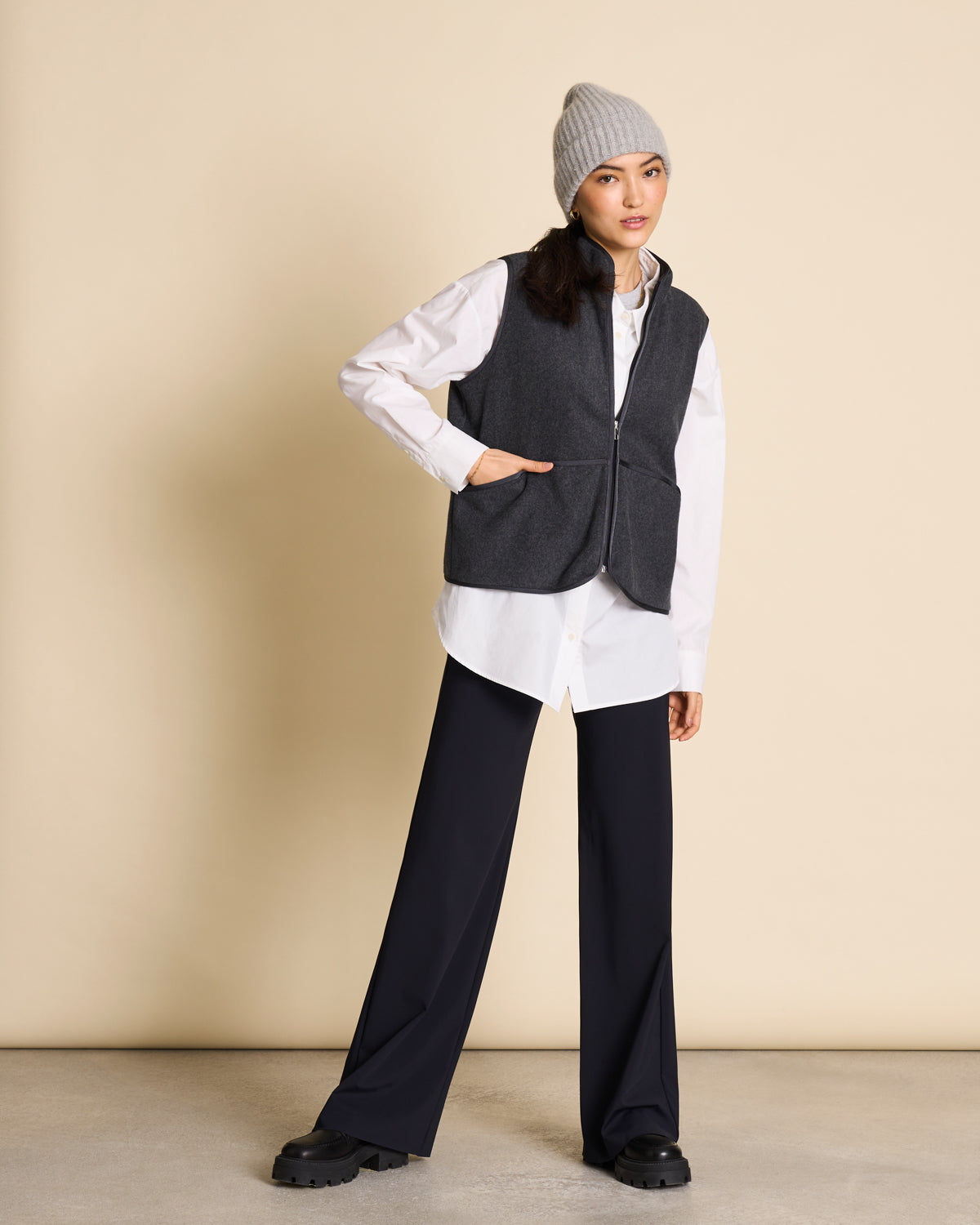 A woman wearing the jan-n-june WOOLY KNIT BEANIE BJON LIGHT GREY, a dark gray vest, white shirt, and black wide-leg pants stands against a beige background, looking at the camera with one hand on her hip.