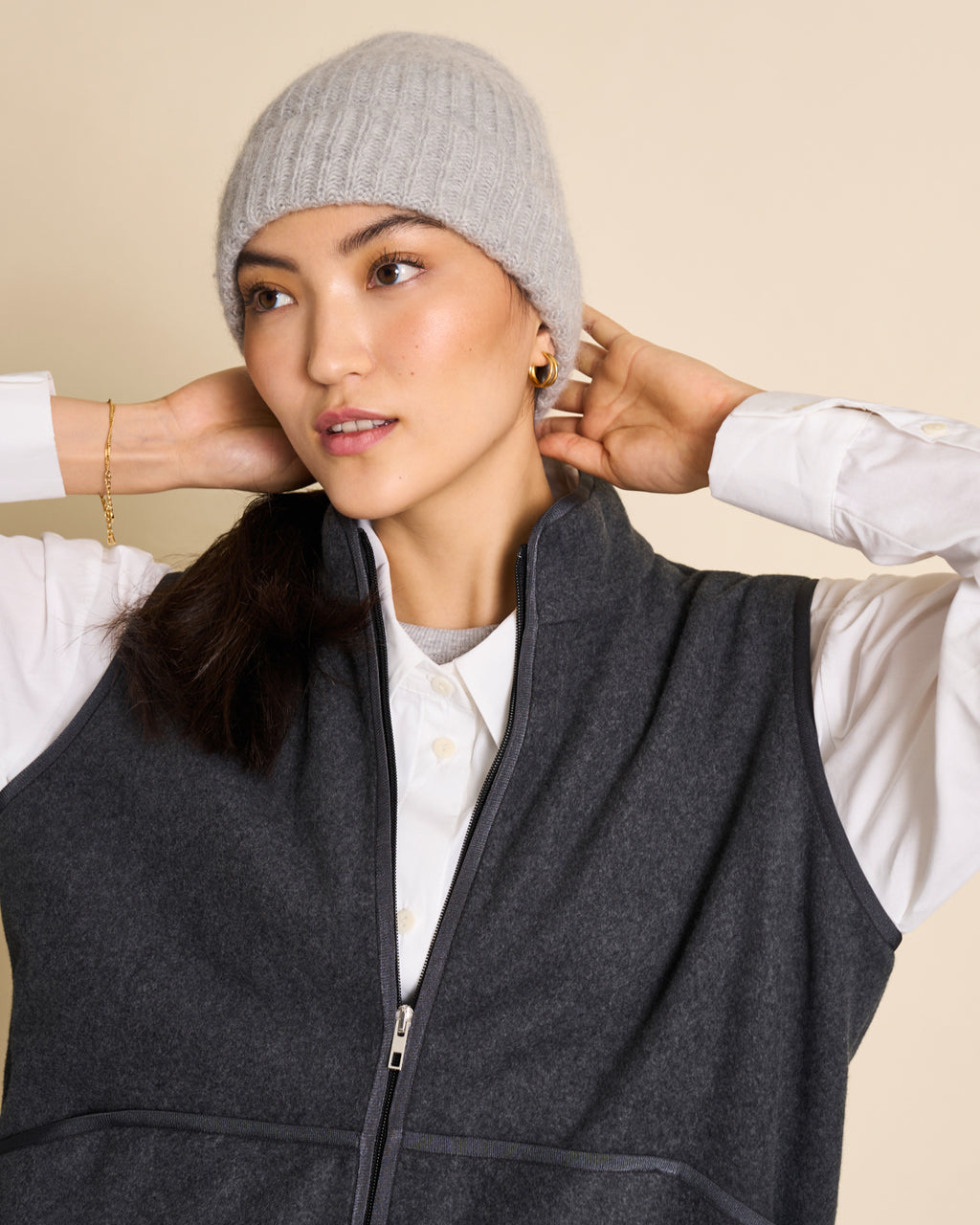 A woman stands against a beige background wearing the jan-n-june WOOLY KNIT BEANIE BJON LIGHT GREY, a white button-up shirt, and a dark gray zip-up vest, with her hands placed behind her head.