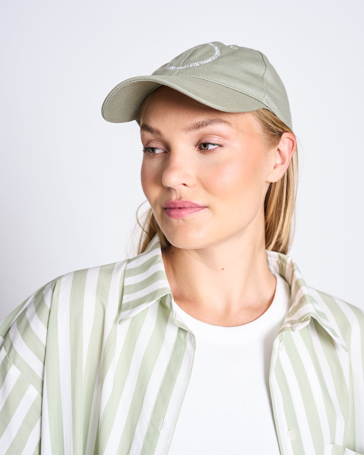 A woman in the jan-n-june CAP FOR THOSE WHO FIX THINGS PISTACHIO GOTS, a timeless organic cotton accessory, and a light green and white striped shirt over a white top, gazes left with a neutral expression against a plain background.