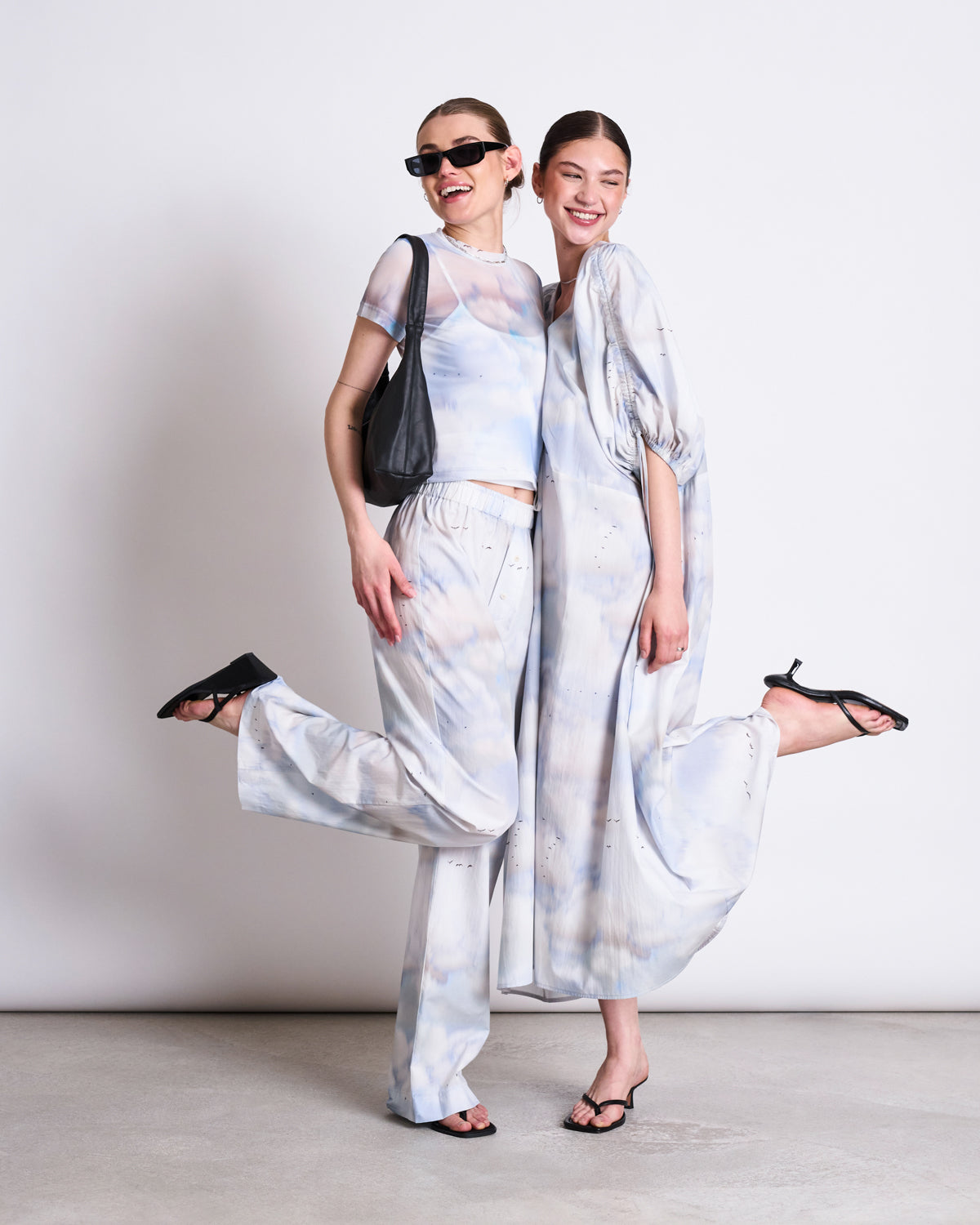 Two women pose playfully on one leg against a white background in jan-n-junes light blue cloud print outfits. One wears sunglasses and pants with a black bag, while the other shows off the flattering MAXI DRESS DANS CLOUD PRINT. Both are smiling.