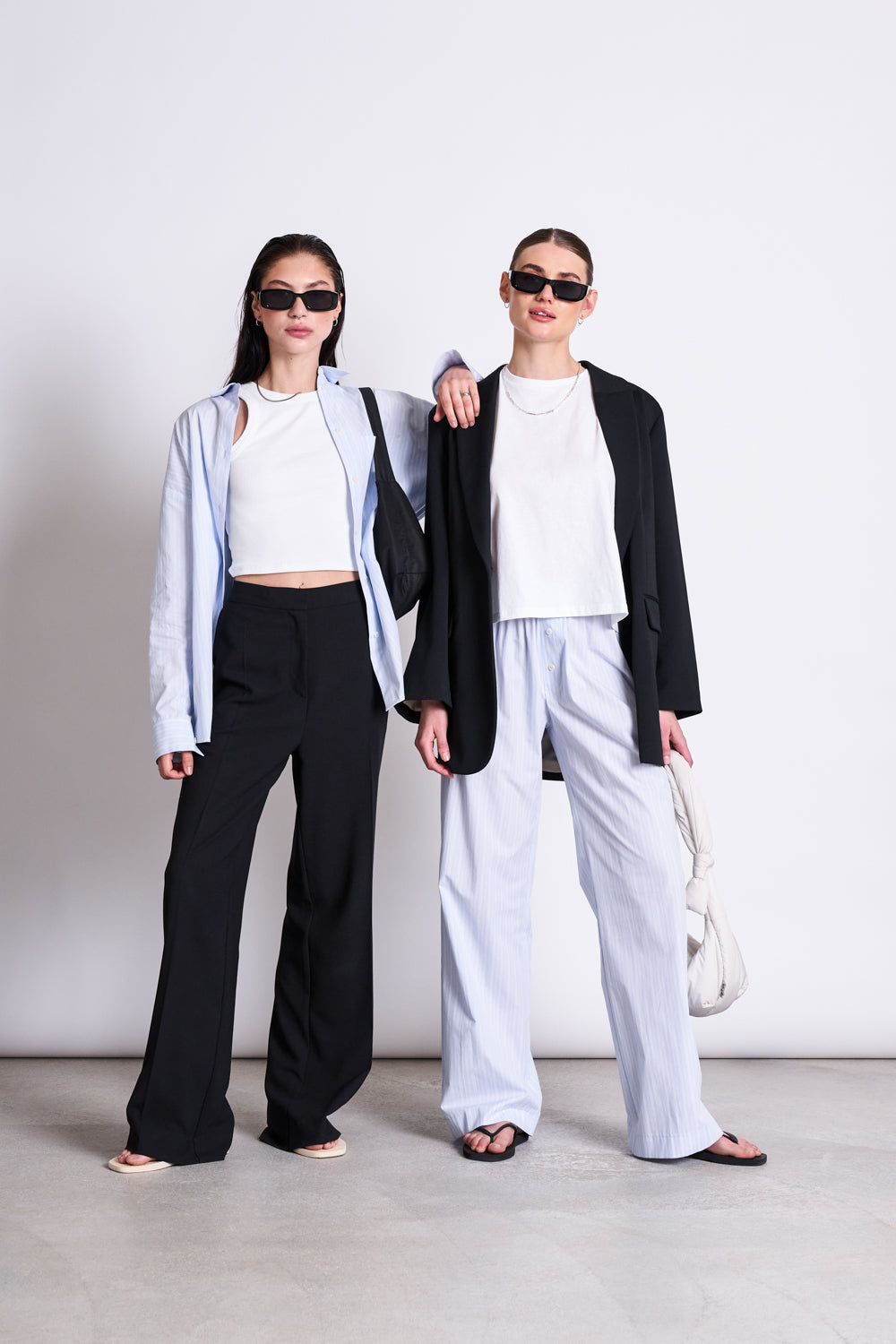 Two women in sunglasses pose confidently against a white background. One wears jan-n-june’s COTTON PANTS SOBY BLUE WHITE STRIPED GOTS with sandals, while the other styles a black blazer with light striped pants and sandals.