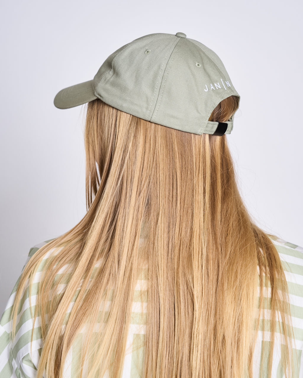 A person with long blonde hair, seen from behind, wears the jan-n-june CAP FOR THOSE WHO FIX THINGS PISTACHIO GOTS—a light green organic cotton baseball cap featuring white text near the back—and a green and white striped shirt.