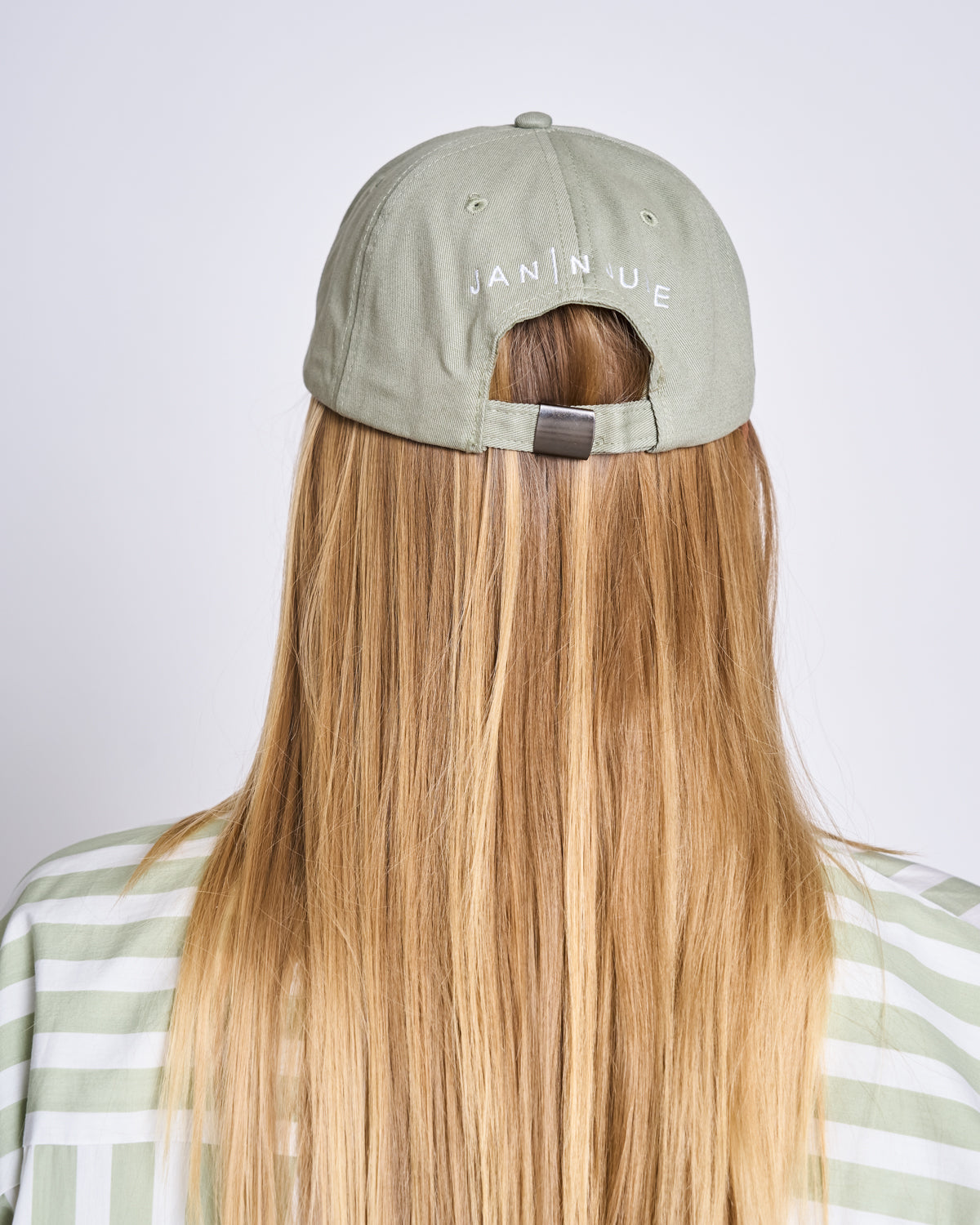 A person with long blonde hair faces away, wearing a pistachio green organic cotton CAP FOR THOSE WHO FIX THINGS by jan-n-june, featuring white JAN|NUE embroidery on the back and a light green and white striped shirt.
