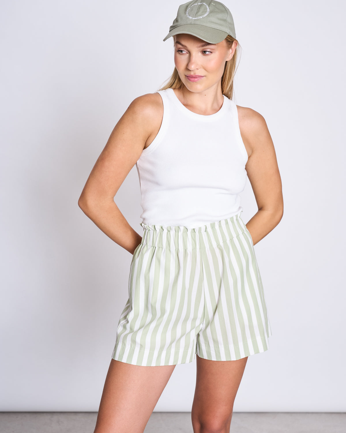 A woman in the CAP FOR THOSE WHO FIX THINGS PISTACHIO GOTS by jan-n-june, a light green cap, wears a white sleeveless tank and high-waisted organic cotton striped shorts, standing with her hands in her pockets against a plain background.