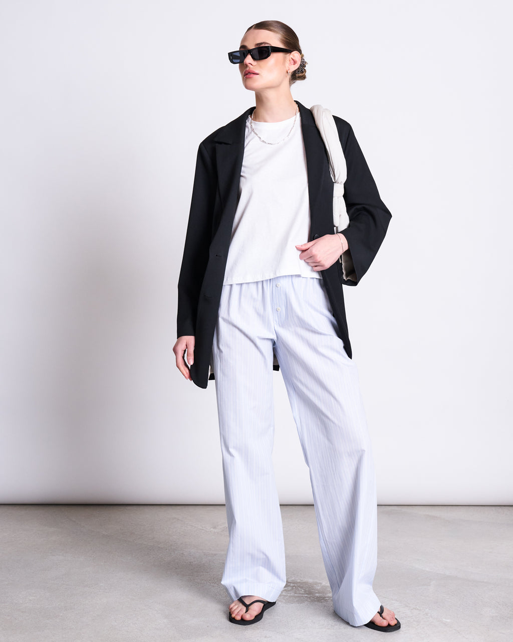 Wearing jan-n-june’s T-SHIRT OVAR WHITE, a black blazer, loose light blue striped pants, sunglasses, black sandals, and carrying a light bag, a person stands against a plain background.