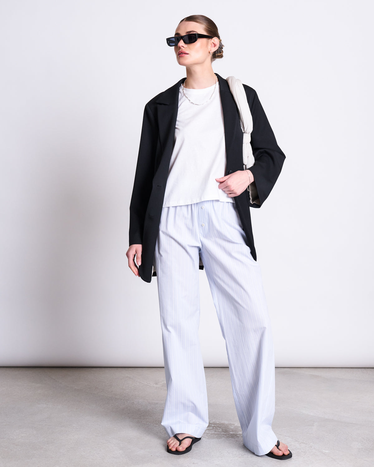 Wearing jan-n-june’s T-SHIRT OVAR WHITE, a black blazer, loose light blue striped pants, sunglasses, black sandals, and carrying a light bag, a person stands against a plain background.