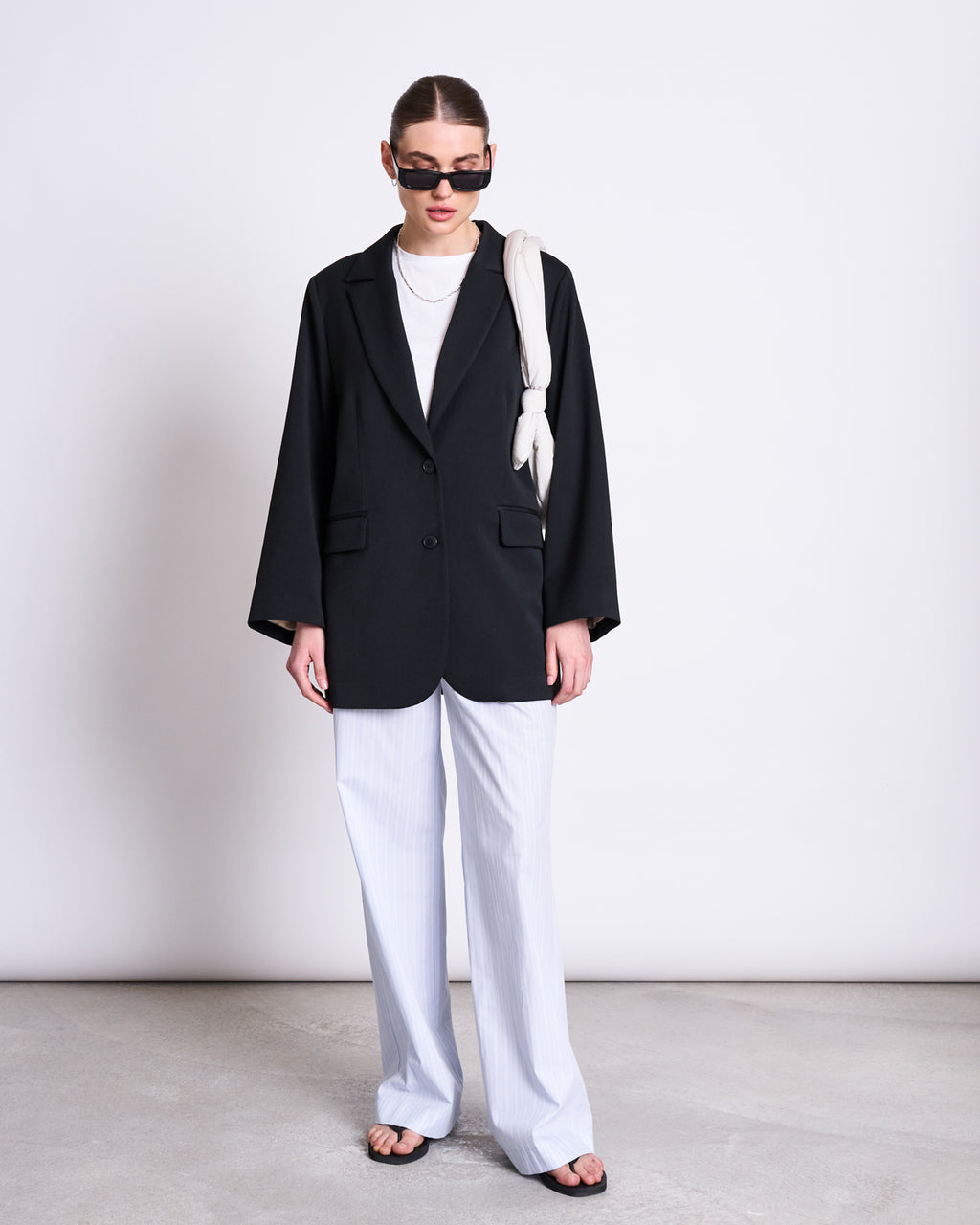 Wearing a black blazer, white shirt, jan-n-june COTTON PANTS SOBY BLUE WHITE STRIPED GOTS, and black sandals, the person stands against a white wall with black sunglasses and a white bag over one shoulder.