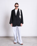 Wearing a black blazer, white shirt, jan-n-june COTTON PANTS SOBY BLUE WHITE STRIPED GOTS, and black sandals, the person stands against a white wall with black sunglasses and a white bag over one shoulder.