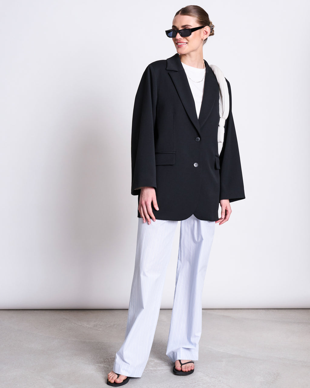 A woman in a black blazer, jan-n-junes COTTON PANTS SOBY BLUE WHITE STRIPED GOTS, black sandals, and sunglasses stands smiling against a plain background with her hair tied back and a white accessory over her shoulder.