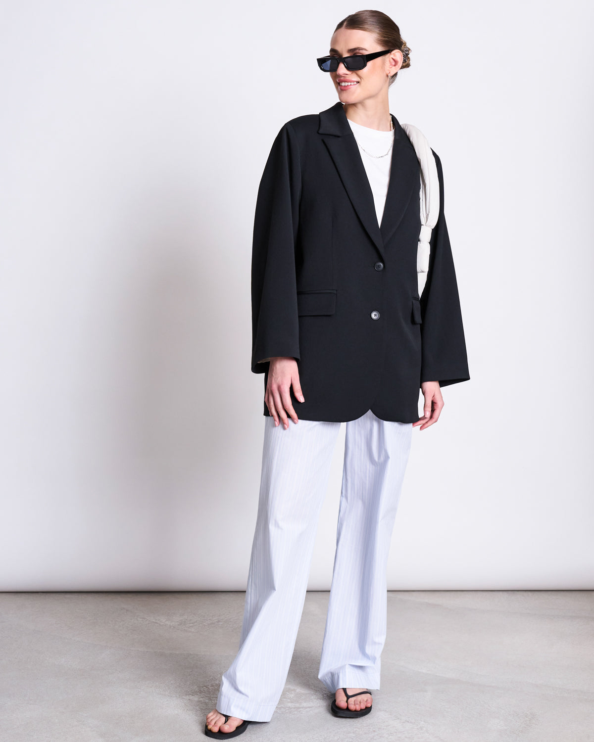 A woman in a black blazer, jan-n-junes COTTON PANTS SOBY BLUE WHITE STRIPED GOTS, black sandals, and sunglasses stands smiling against a plain background with her hair tied back and a white accessory over her shoulder.