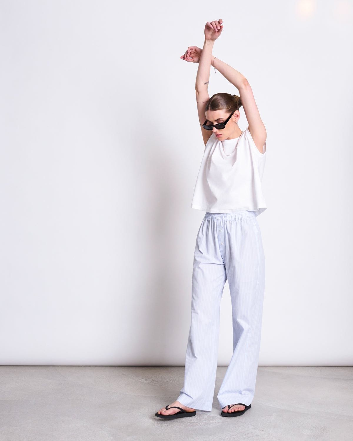 A woman in sunglasses, a sleeveless white top, jan-n-june’s COTTON PANTS SOBY BLUE WHITE STRIPED GOTS, and black sandals stands against a plain white wall with her arms raised above her head.