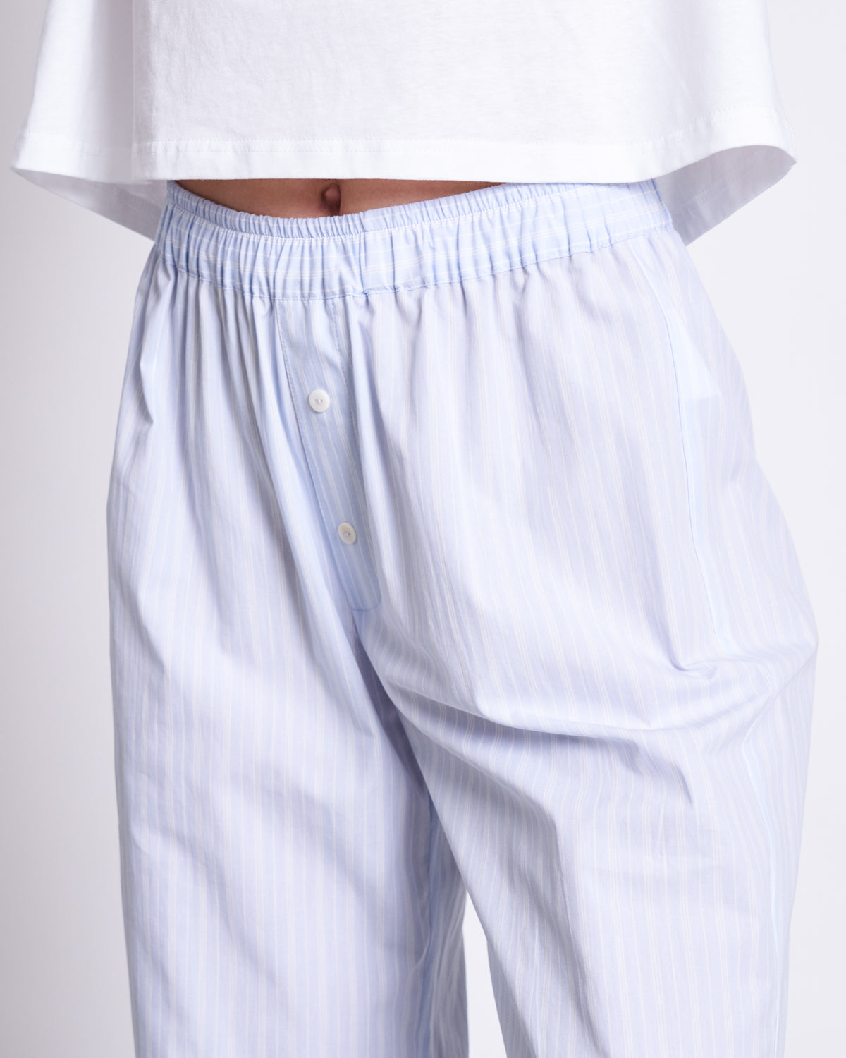 A person wears jan-n-junes COTTON PANTS SOBY BLUE WHITE STRIPED GOTS—comfortable, organic cotton pyjama pants with light blue and white stripes, a white elastic waistband, and buttons—paired with a cropped white shirt.