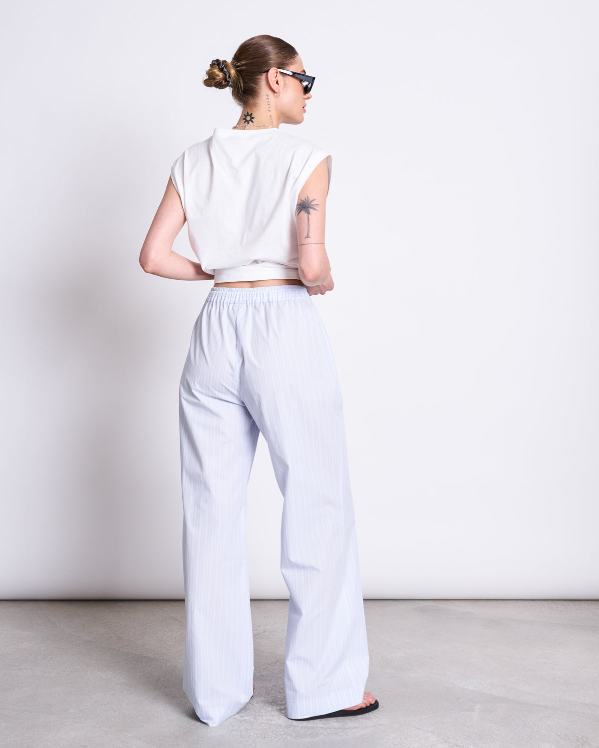 A woman with a bun and sunglasses stands in a minimal studio, showing tattoos on her neck and left arm while wearing a white sleeveless crop top and jan-n-junes COTTON PANTS SOBY BLUE WHITE STRIPED GOTS.