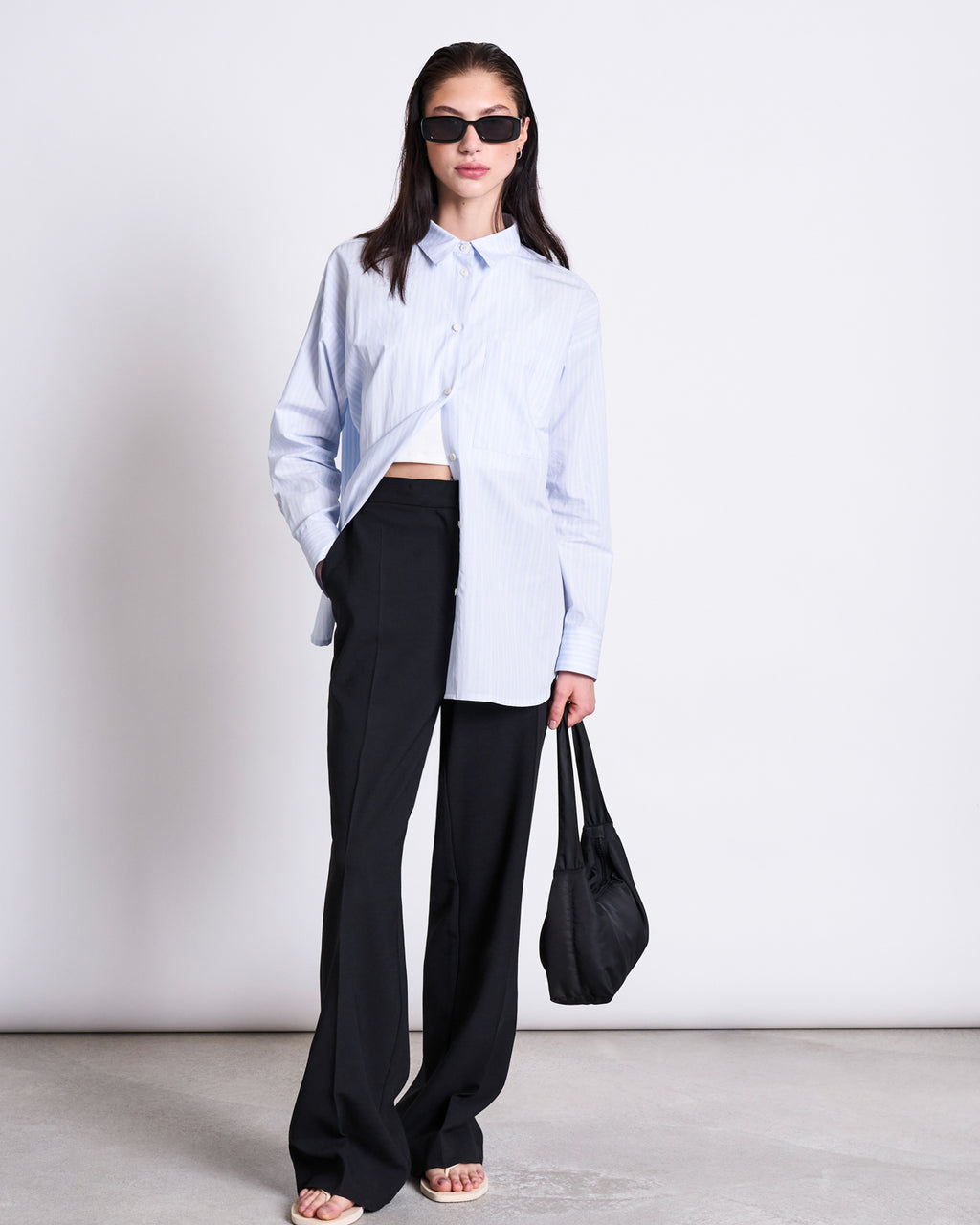 A woman stands against a white background wearing jan-n-junes ECOVERO™ WIDE PANTS CORALIE BLACK, paired with black sunglasses, a light blue button-up shirt (half tucked), white sandals, and holding a black handbag.
