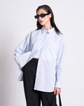A person with long dark hair, black sunglasses, and black pants poses against a white background wearing the jan-n-june OVERSIZED SHIRT MAMRO in blue white striped GOTS, with one hand in their pocket.