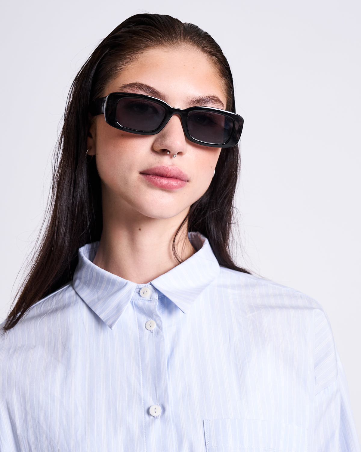 A young woman with straight brown hair wears black rectangular sunglasses and the jan-n-june OVERSIZED SHIRT MAMRO BLUE WHITE STRIPED GOTS, standing against a plain white background.