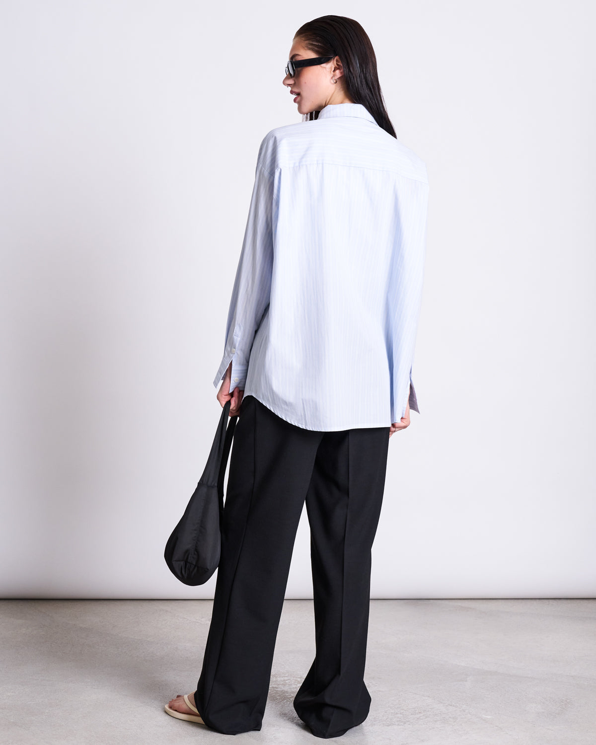 A woman stands with her back turned, wearing the jan-n-june OVERSIZED SHIRT MAMRO BLUE WHITE STRIPED GOTS, paired with loose black trousers, sandals, sunglasses, and carrying a black bag against a plain white background.