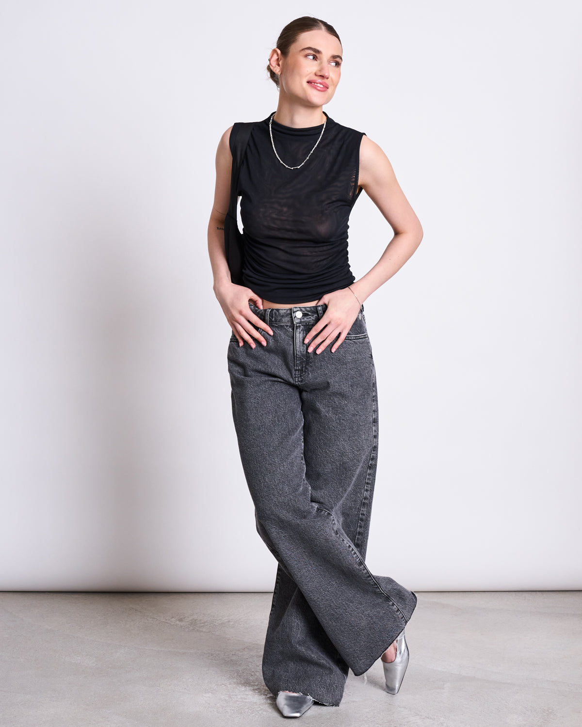 A woman smiles and looks to the side, wearing the jan-n-june RUFFLE MESH TOP LAGUNA BLACK with high-waisted loose gray jeans, silver shoes, and a silver necklace against a plain light background. She stands with her legs crossed.