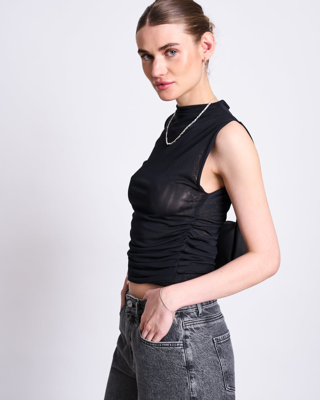 A woman with light skin and brown hair tied back wears the jan-n-june RUFFLE MESH TOP LAGUNA BLACK, a sleeveless sheer black top with a high neckline, paired with gray jeans. She stands against a white background, hand in pocket, looking at the camera.